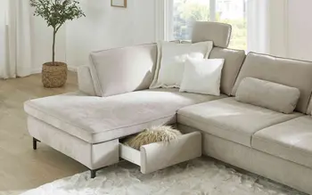 Frau genießt weiche Stoffe in verschiedenen Farben, von Beige bis Senfgelb. Rechts ein komfortables Ecksofa mit Stauraum-Schublade und weißen Kissen im hellen Wohnzimmer – perfekte Kombination aus Design und Funktionalität.